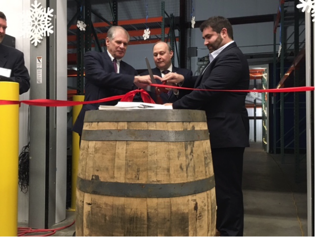 Image of three men cutting a ribbon over a giant barrel. 